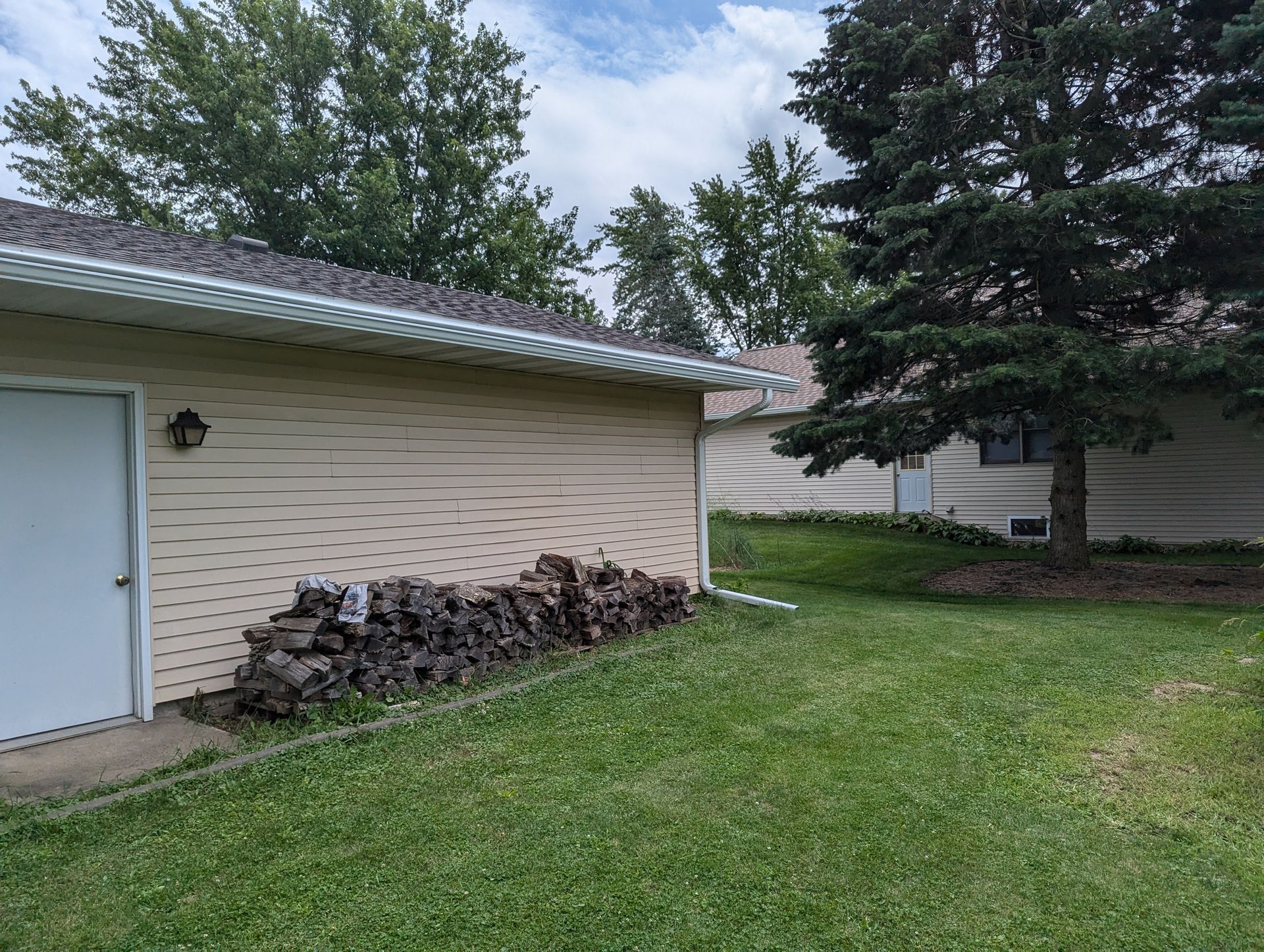 Tan building with a white door, a pile of wood, and green grass. Trees in the background.