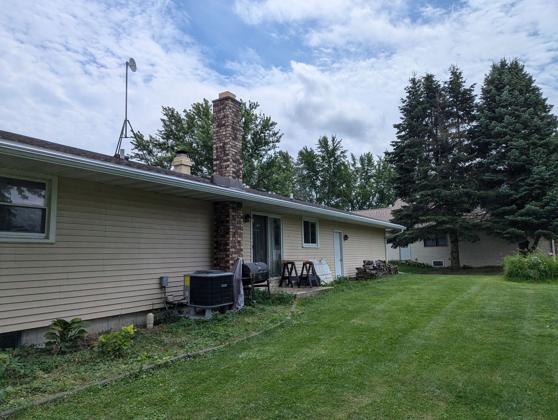Tan house with a chimney, air conditioner, and green lawn under a cloudy sky.