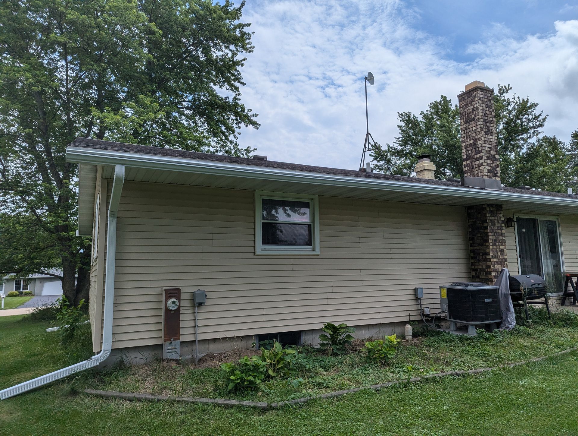 Beige-sided house with a window, chimney, and an air conditioning unit; green lawn under a cloudy sky.