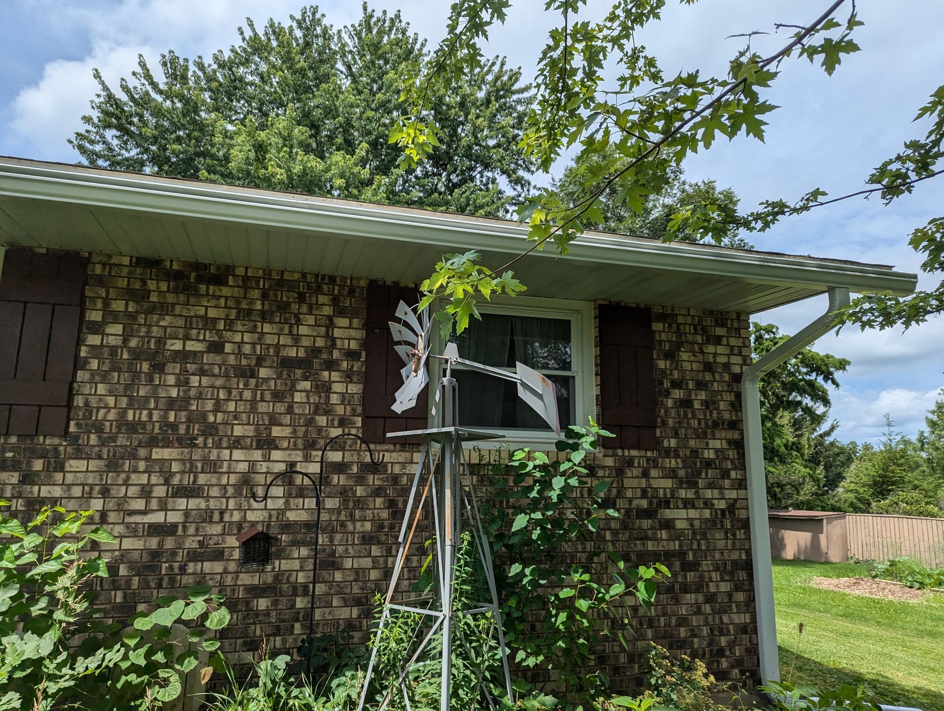 Brick building with a decorative windmill in front of a window, surrounded by greenery. Brown shutters and white gutters.
