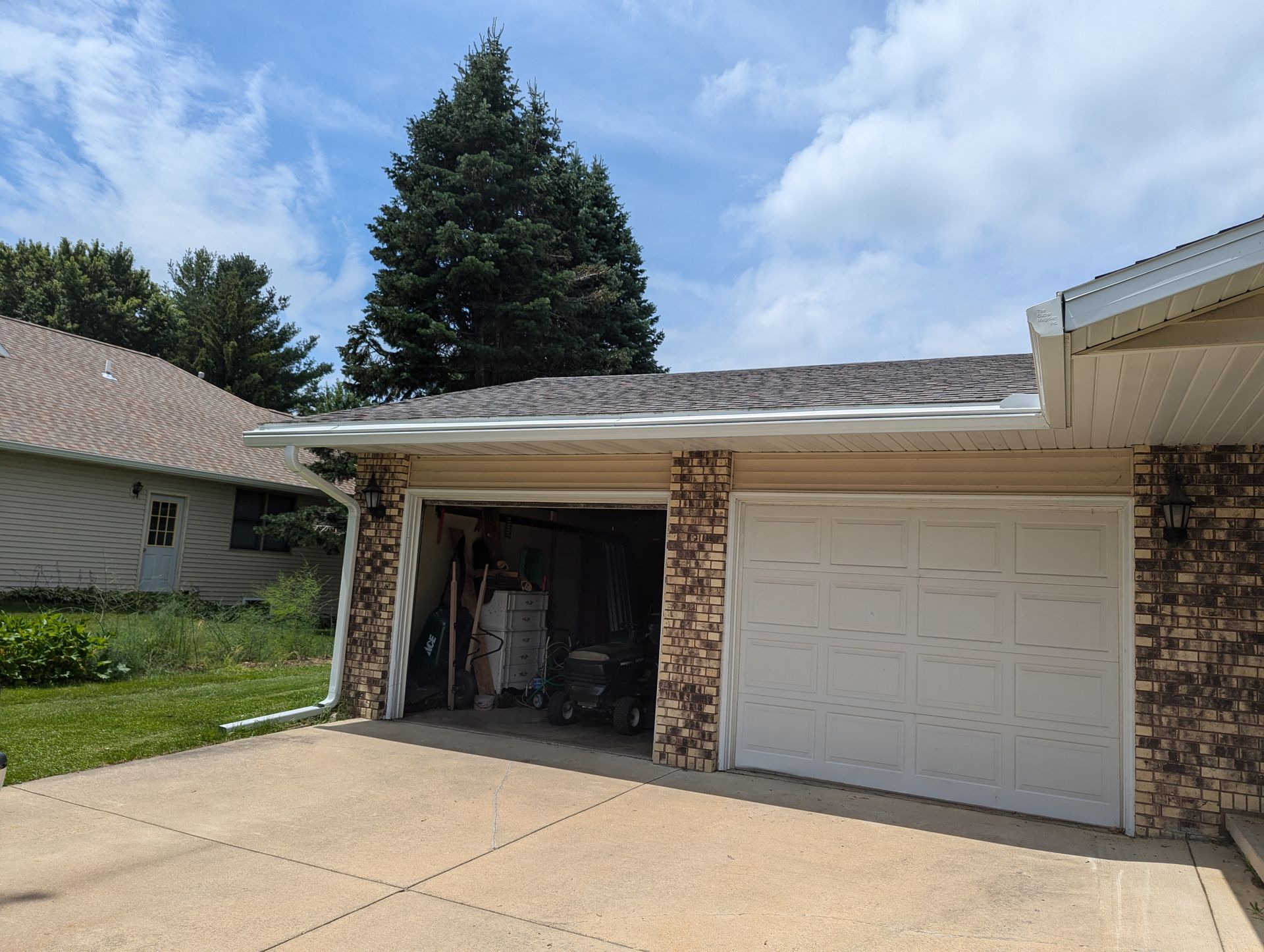 Garage with closed white door, open bay with items, brick facade, concrete driveway, green tree, and cloudy sky.
