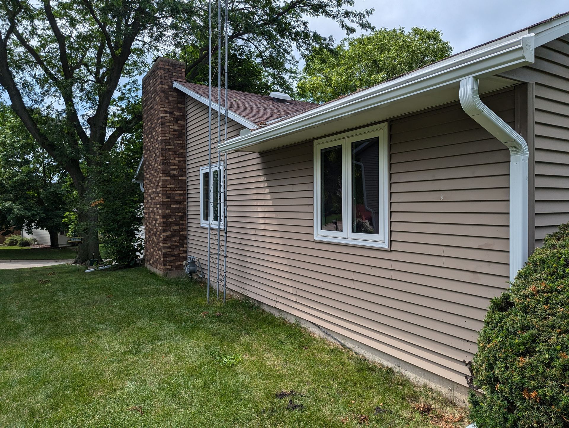 Tan-sided house with white trim, chimney, and a grassy lawn under a cloudy sky.