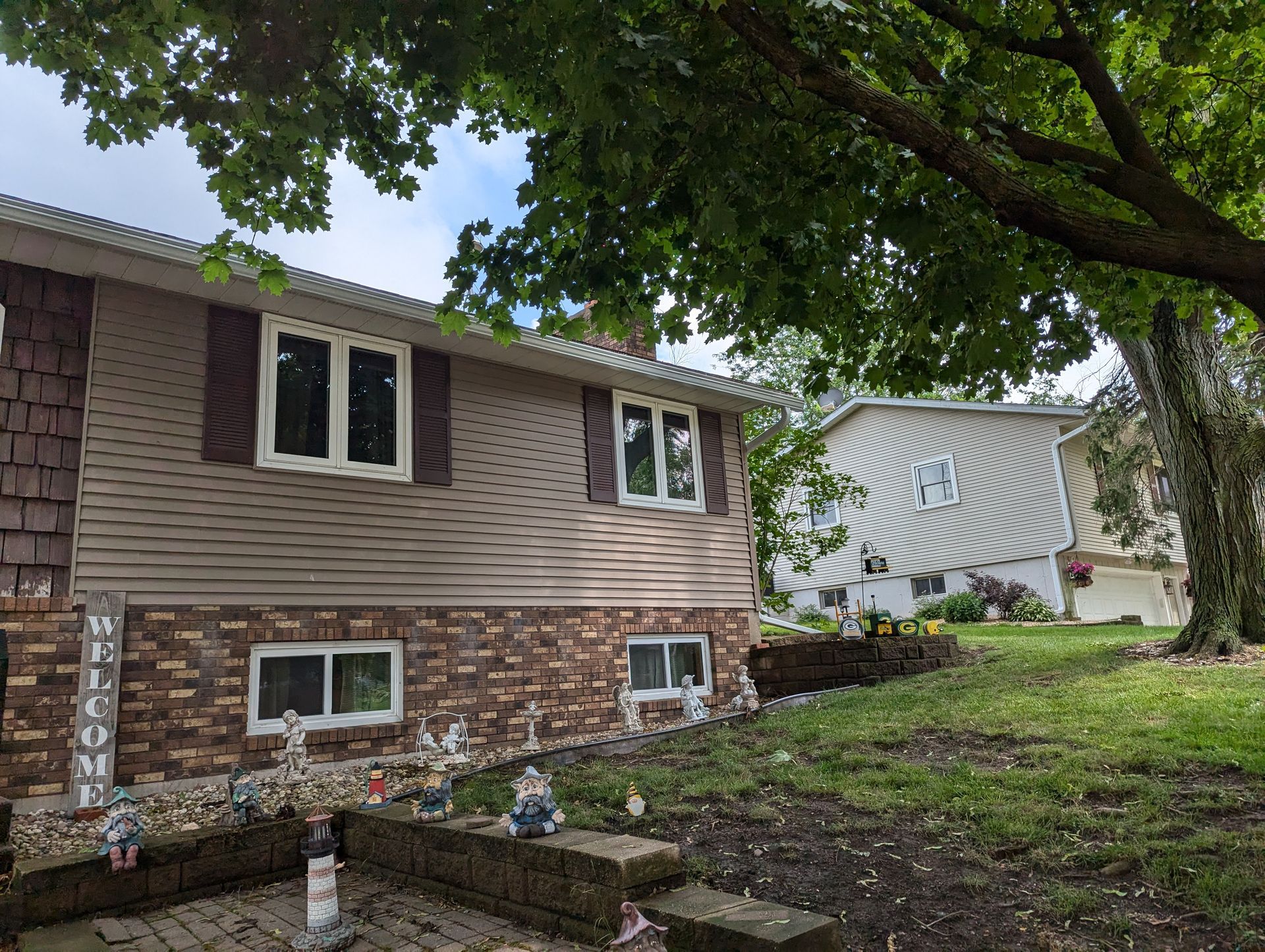 Tan and brown house with stone and siding, surrounded by trees and lawn.