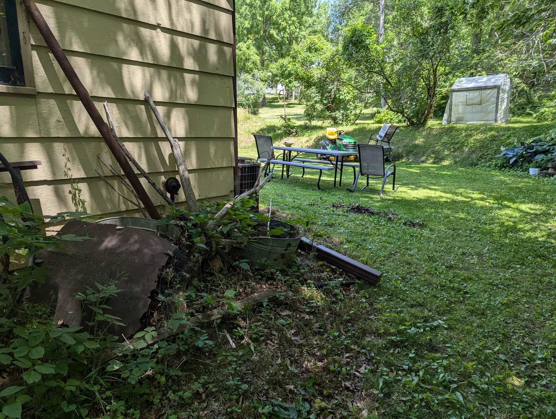 Lush backyard with picnic table, chairs, and overgrown plants next to a house with an attached metal structure.