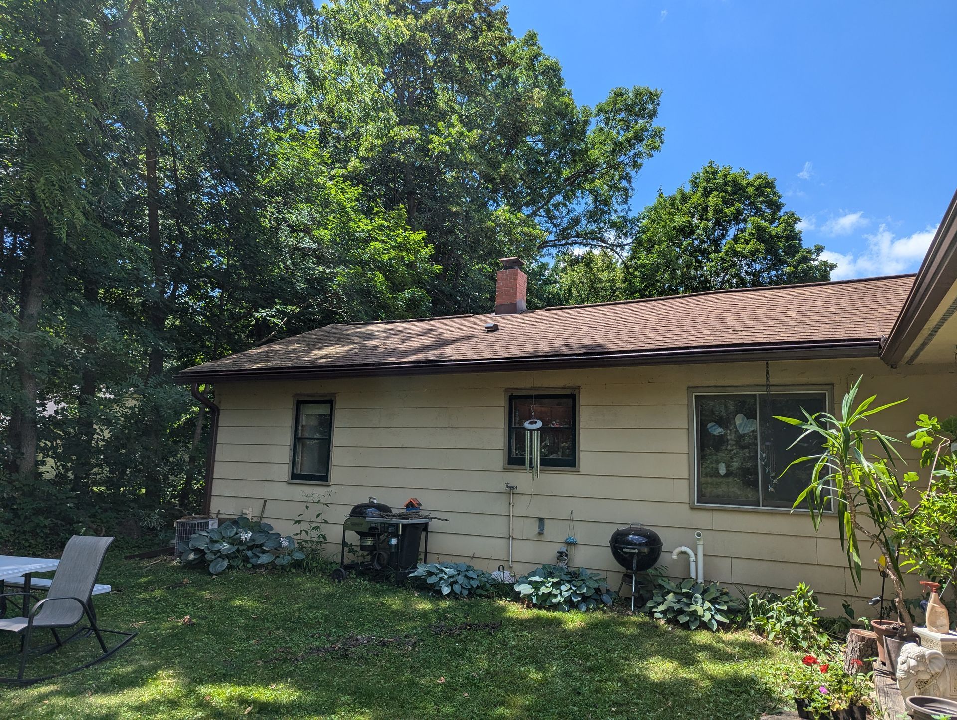 Beige house with a brown roof and chimney, backyard with trees and a grill.