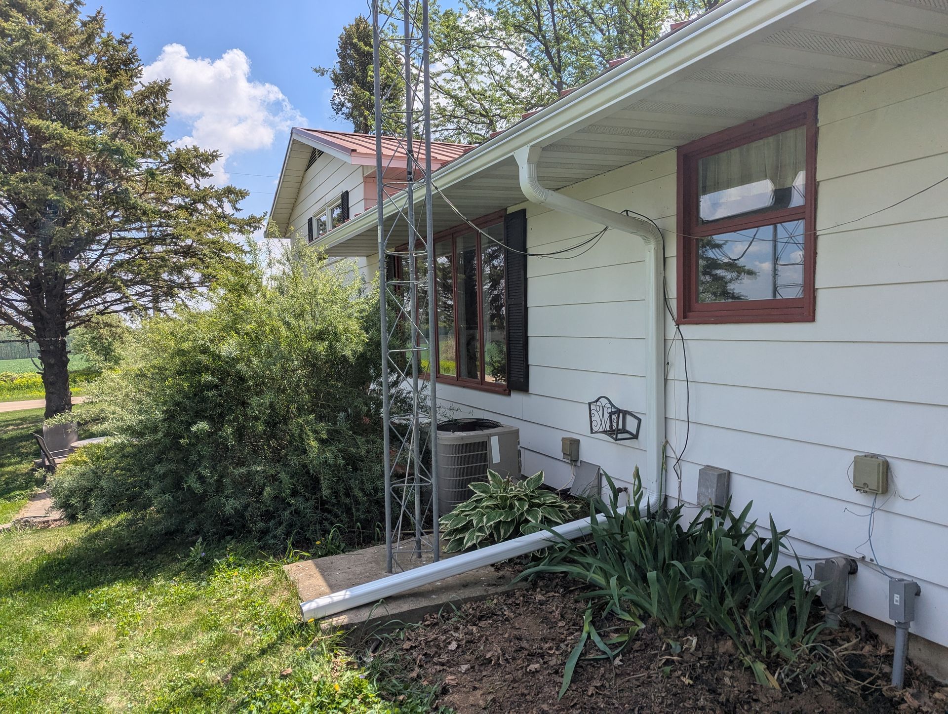 White house with brown trim and windows, next to a metal tower and some shrubbery on a sunny day.