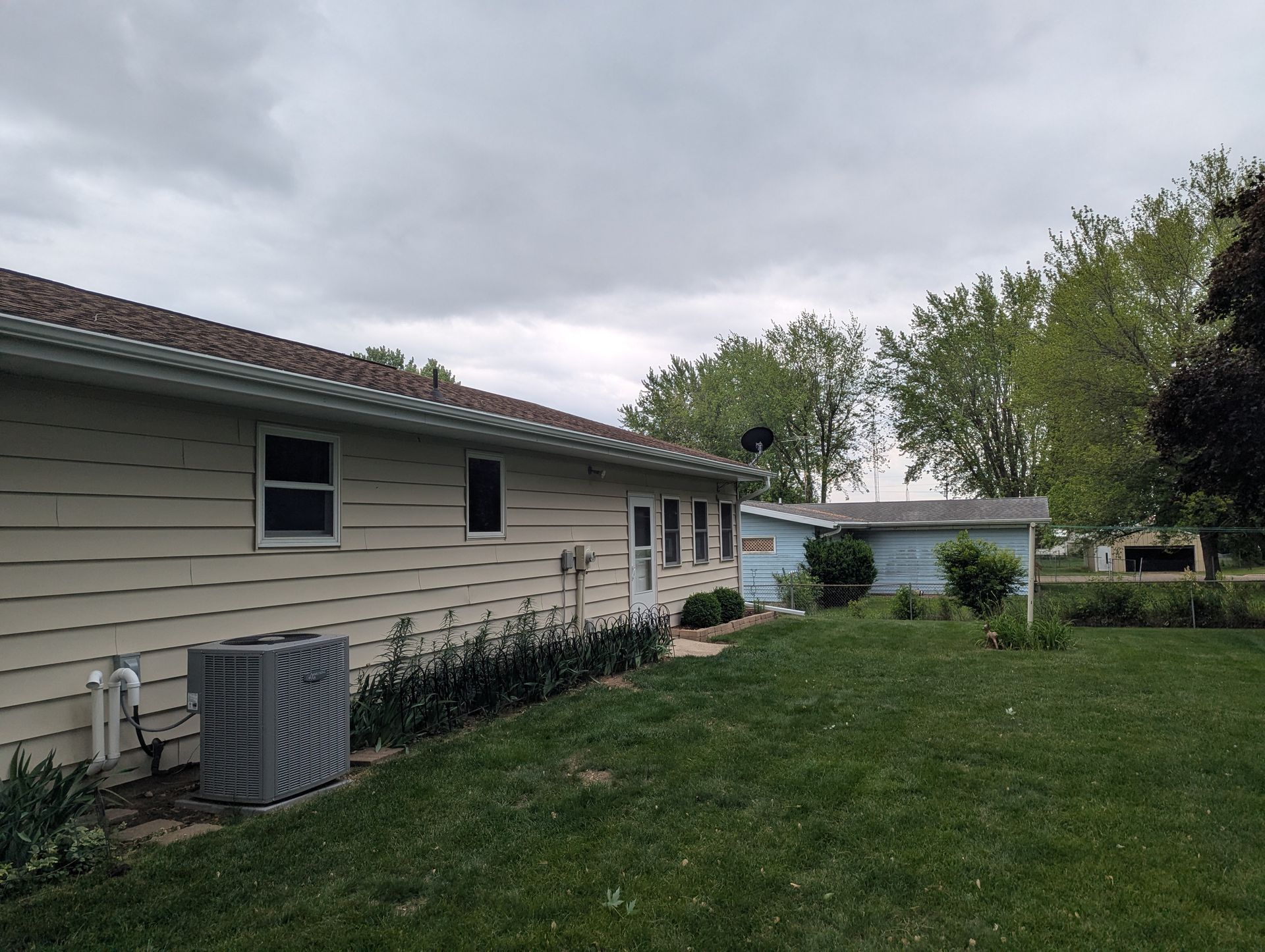 Side view of a house with beige siding, dark roof, air conditioning unit, and green lawn under a cloudy sky.
