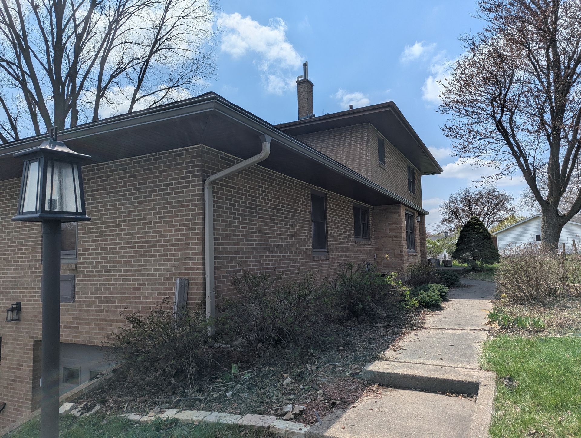 Brick house with a walkway, surrounded by greenery and trees, under a partly cloudy sky.