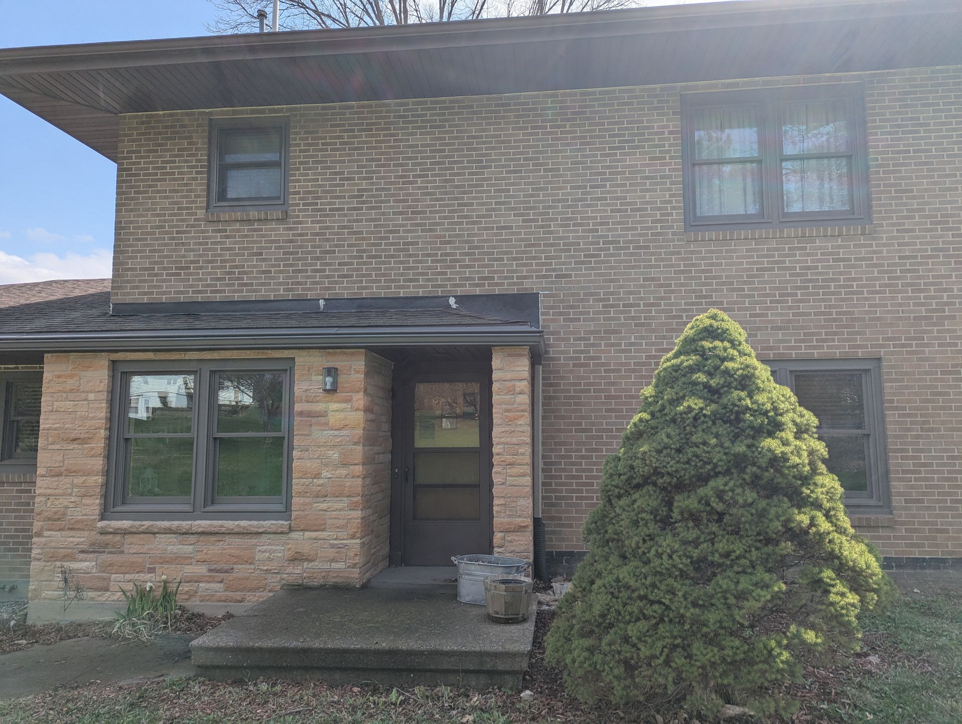 Brick two-story house with dark trim and front door under a small overhang. A green bush is on the right.
