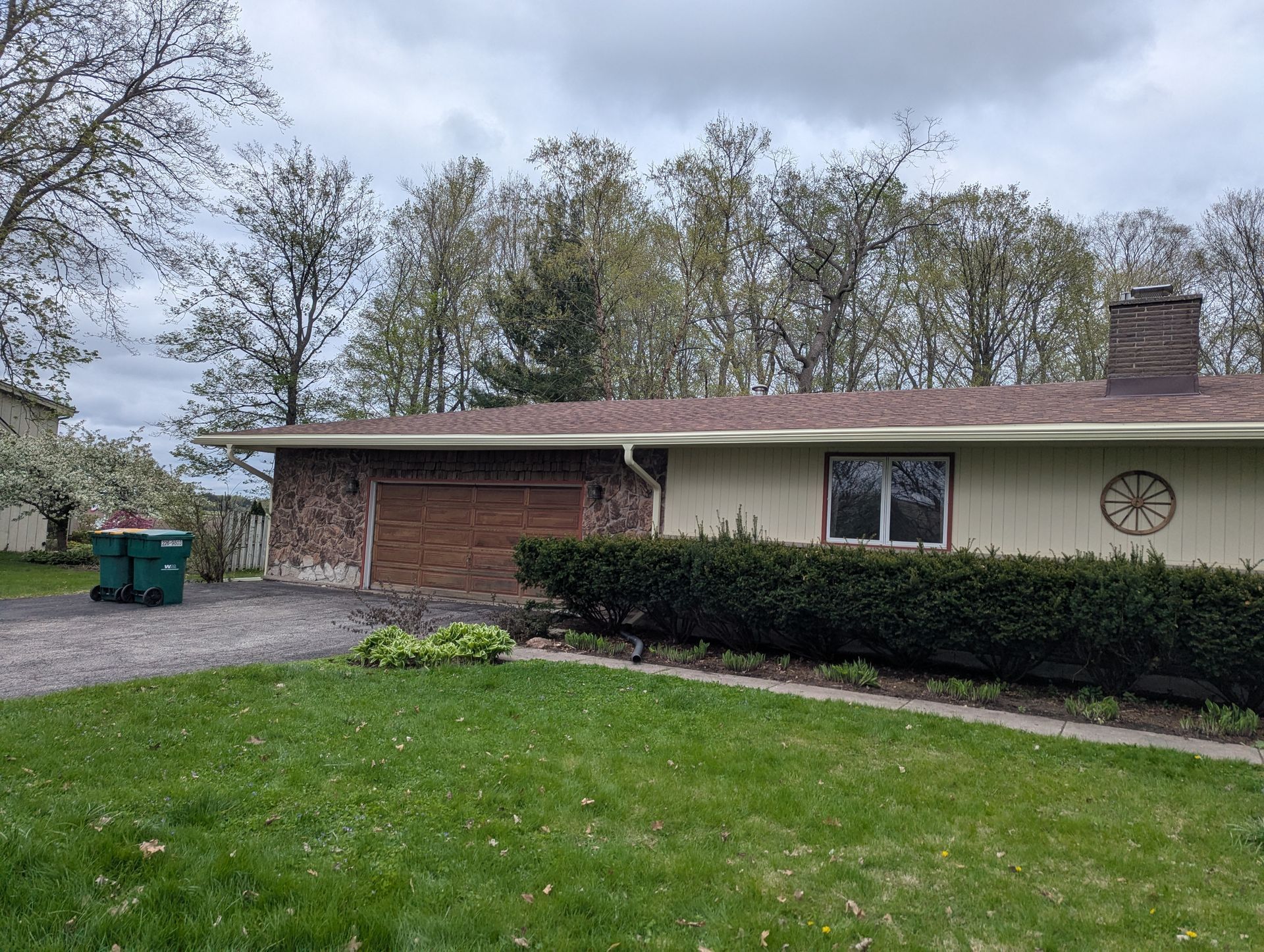 House with brick garage, tan siding, brown roof, and green lawn under a cloudy sky.