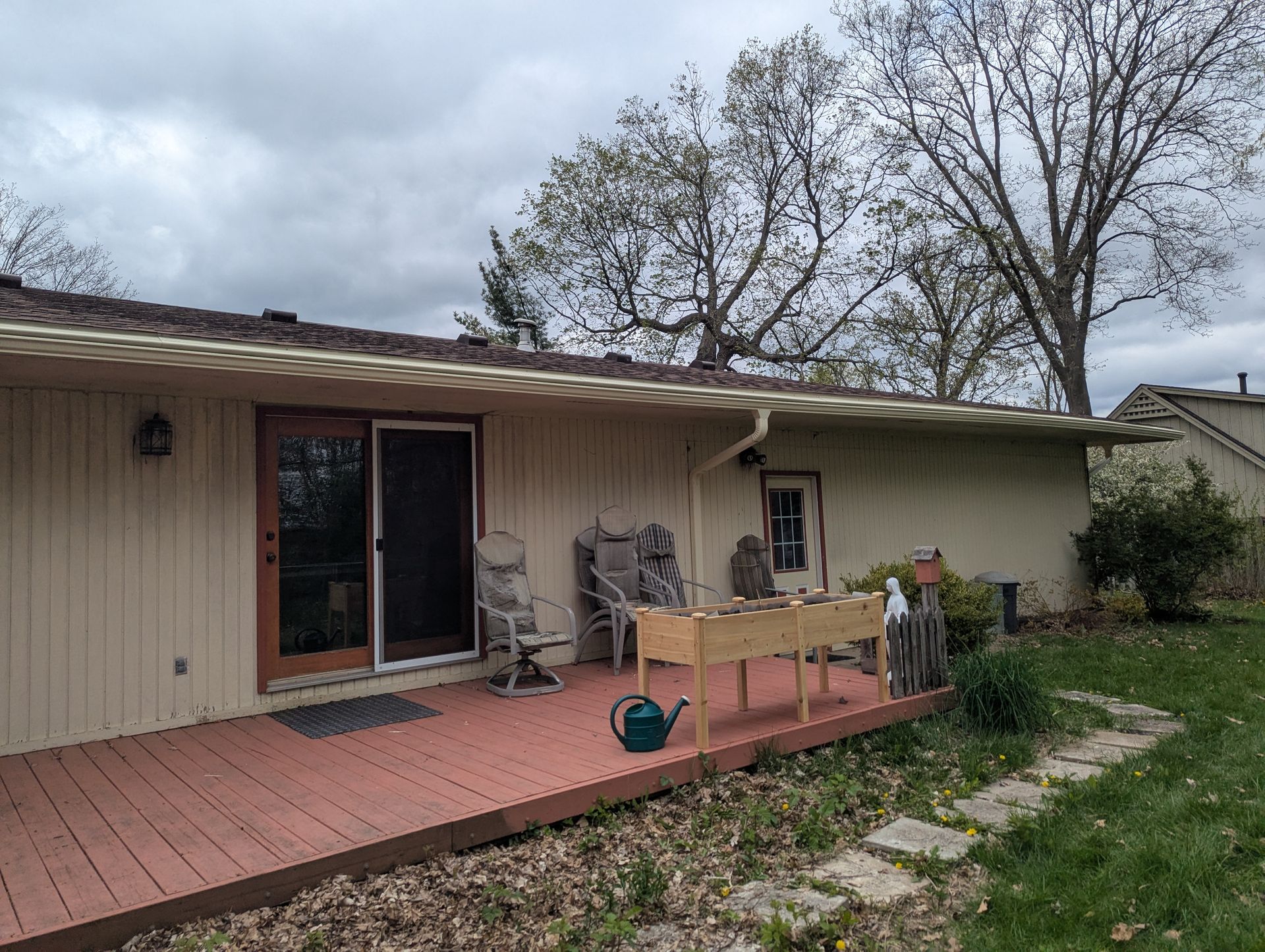 Backyard deck with a wooden planter, chairs, and sliding glass door. Overcast sky.
