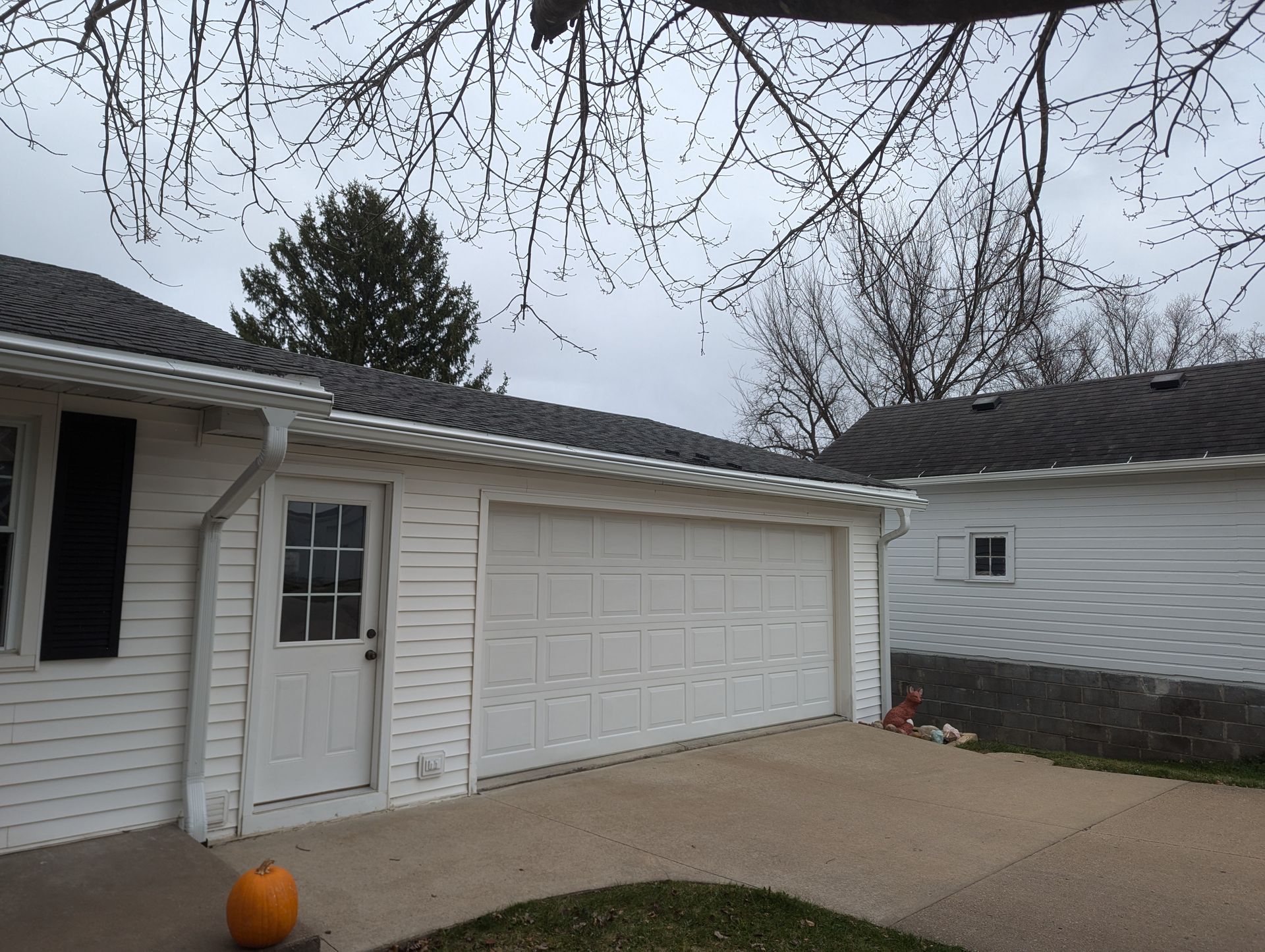 White garage with a door and closed garage door, white house siding, grey roof, and a pumpkin.