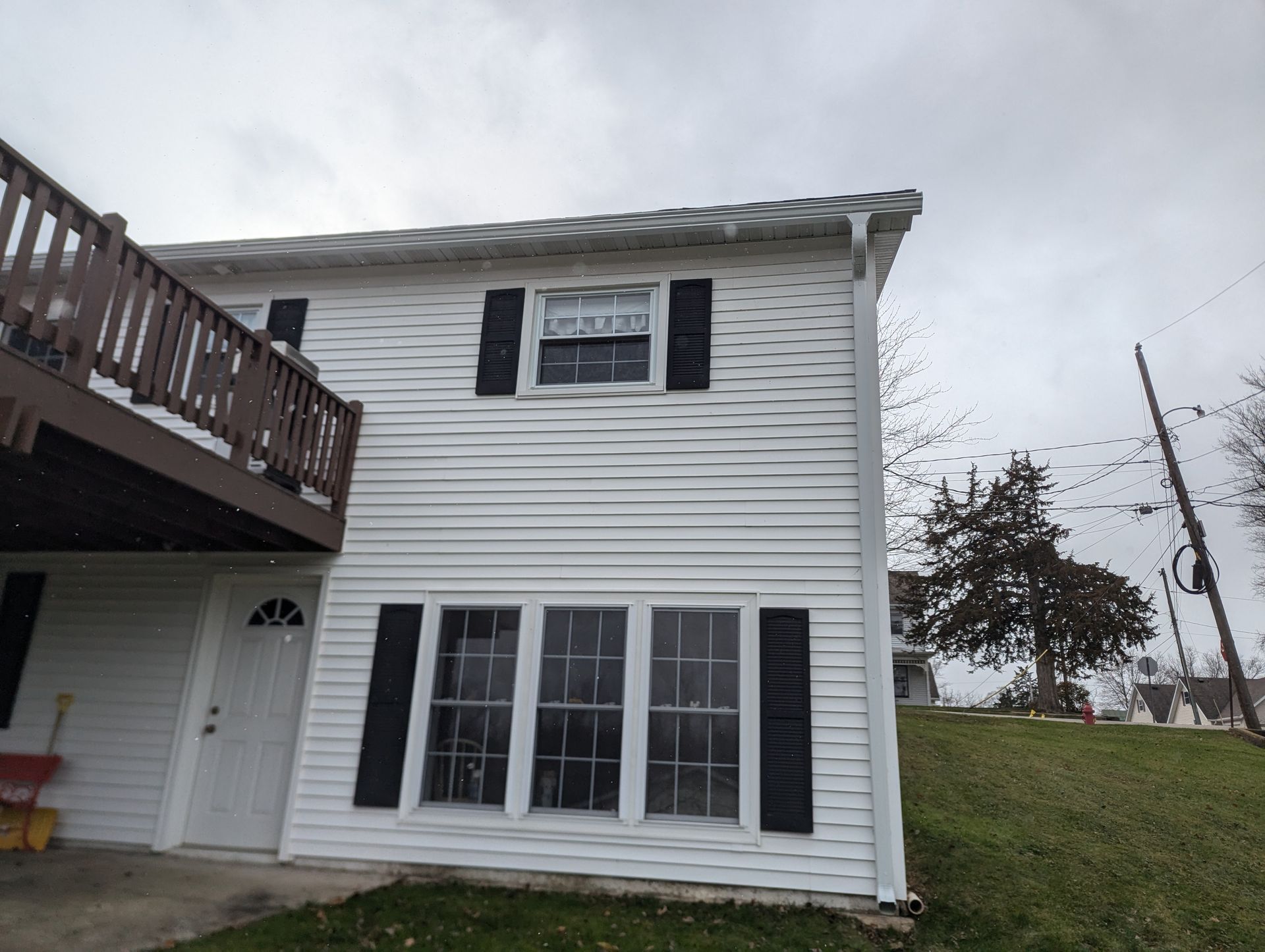 White two-story building with black shutters and a brown balcony against a cloudy sky.
