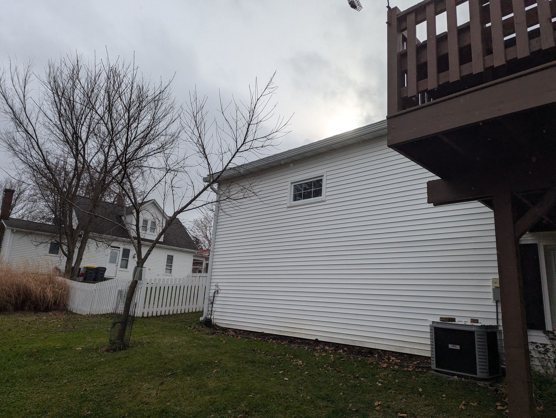 Backyard view with a house, bare tree, fence, and a wooden deck under a cloudy sky.