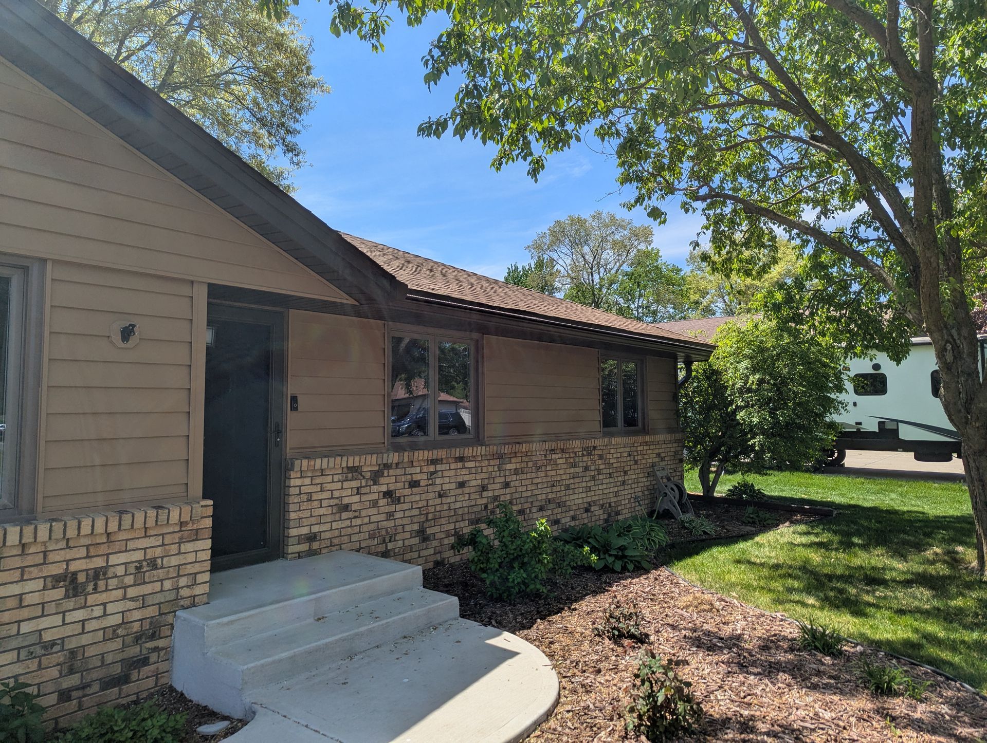 Tan house with brick and a dark roof, surrounded by greenery and trees.