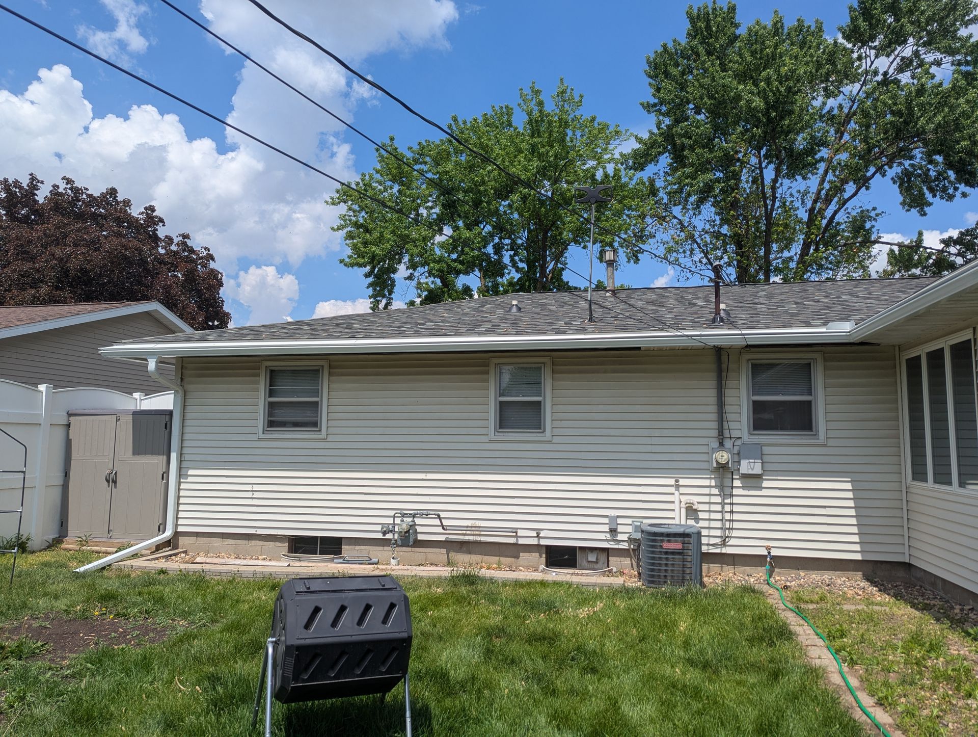 Backyard of a house with white siding, windows, and a gray roof. Green grass and trees are in the background.