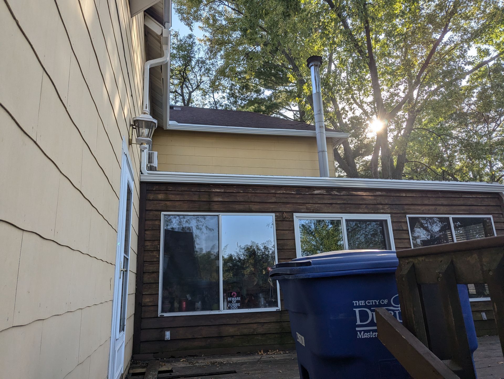 Exterior of a house with yellow and brown siding, blue trash can, and tall chimney.