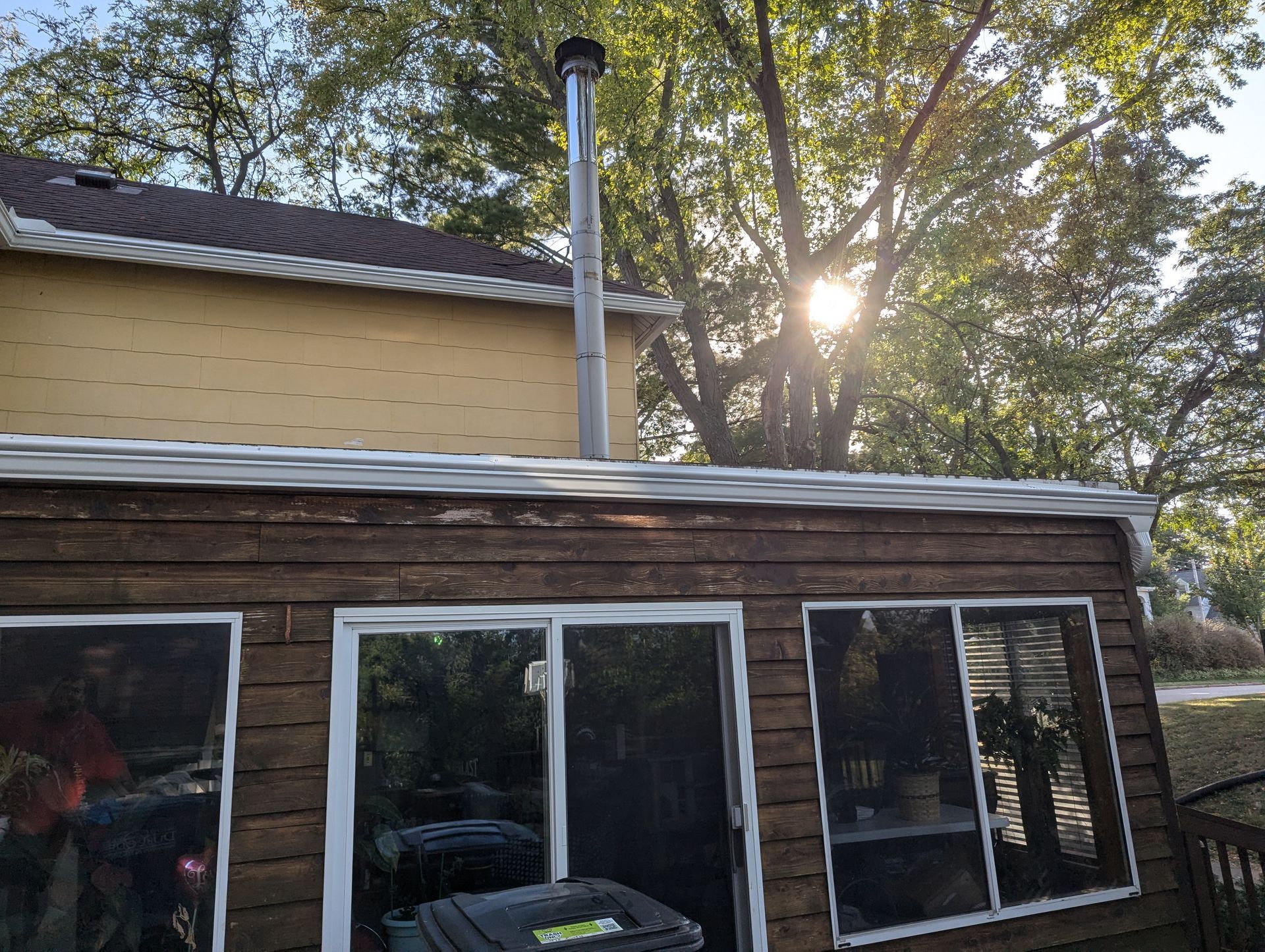 Exterior view of a house with a tall metal chimney. The sun shines through trees in the background.