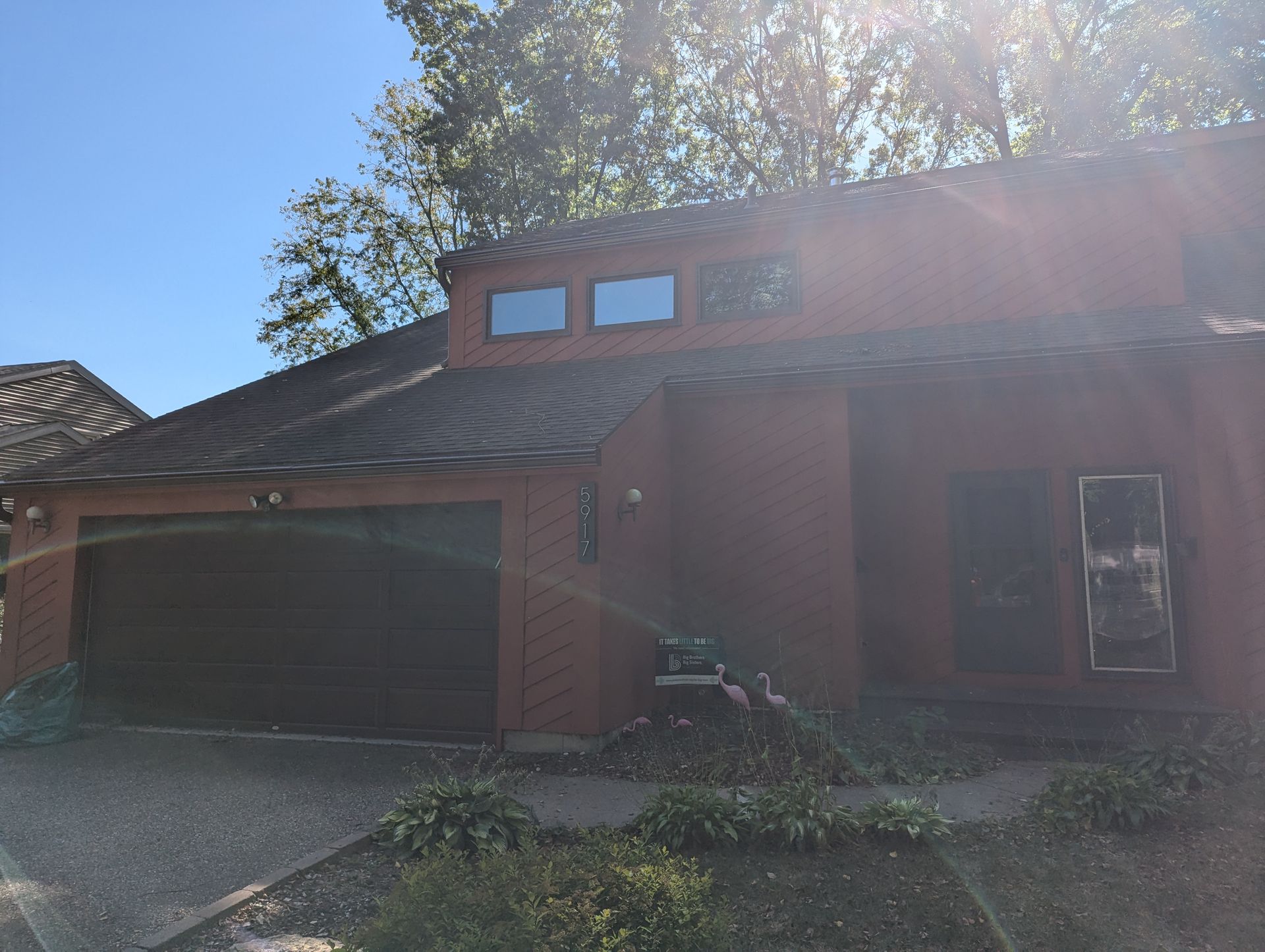 Red-brown house with a dark brown garage door, windows, and bushes in front, against a blue sky.