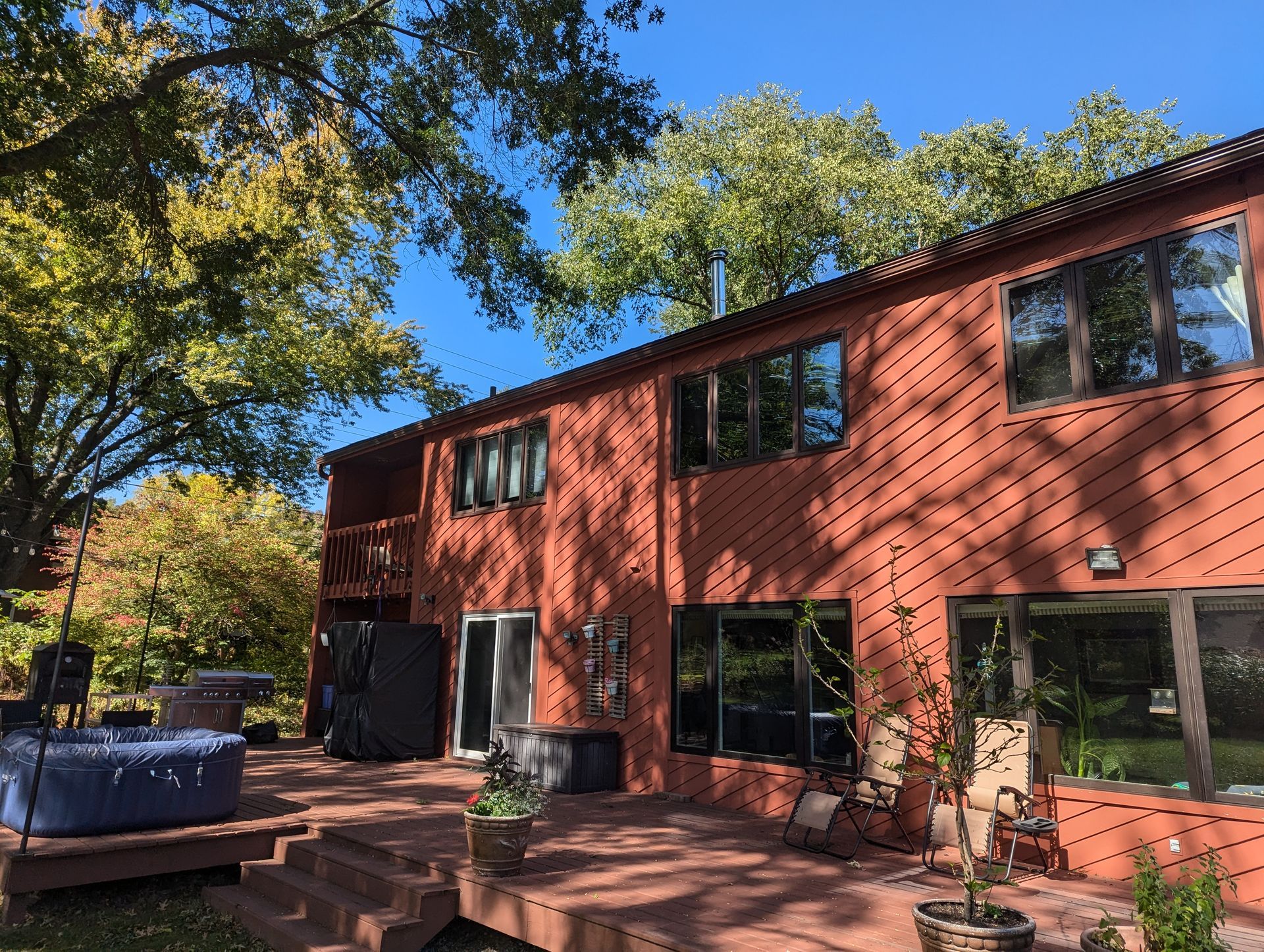 Red-sided two-story house with large windows, wooden deck, and trees under a blue sky.