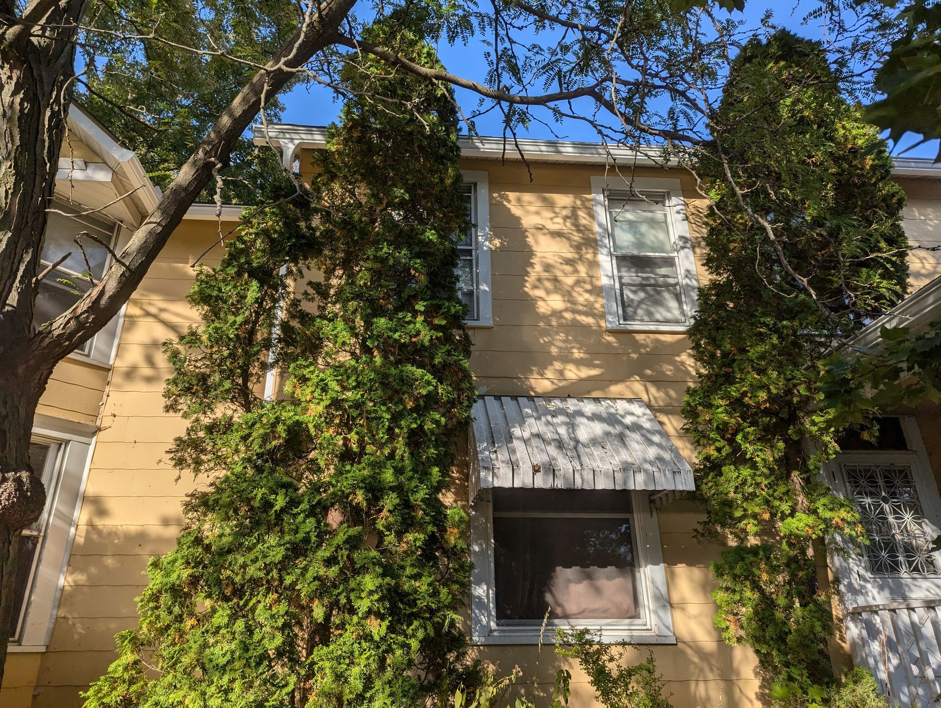 Yellow house exterior with green trees, white window frames, and an awning.