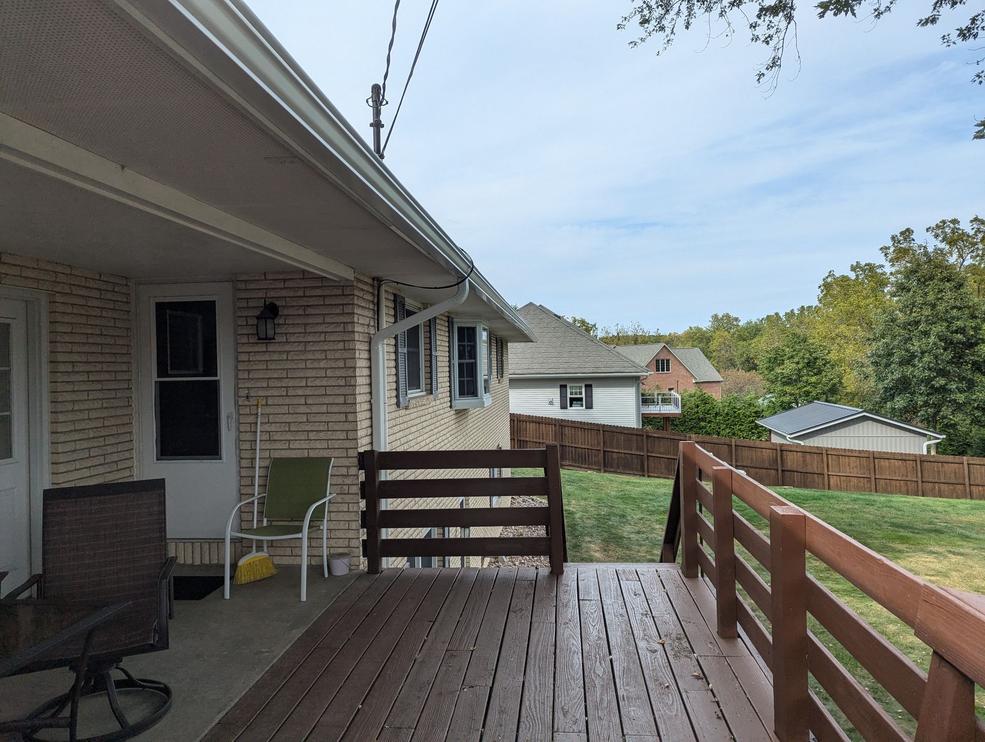 Backyard deck with brown railing, leading to a lawn with a fence. A brick house is in the background.