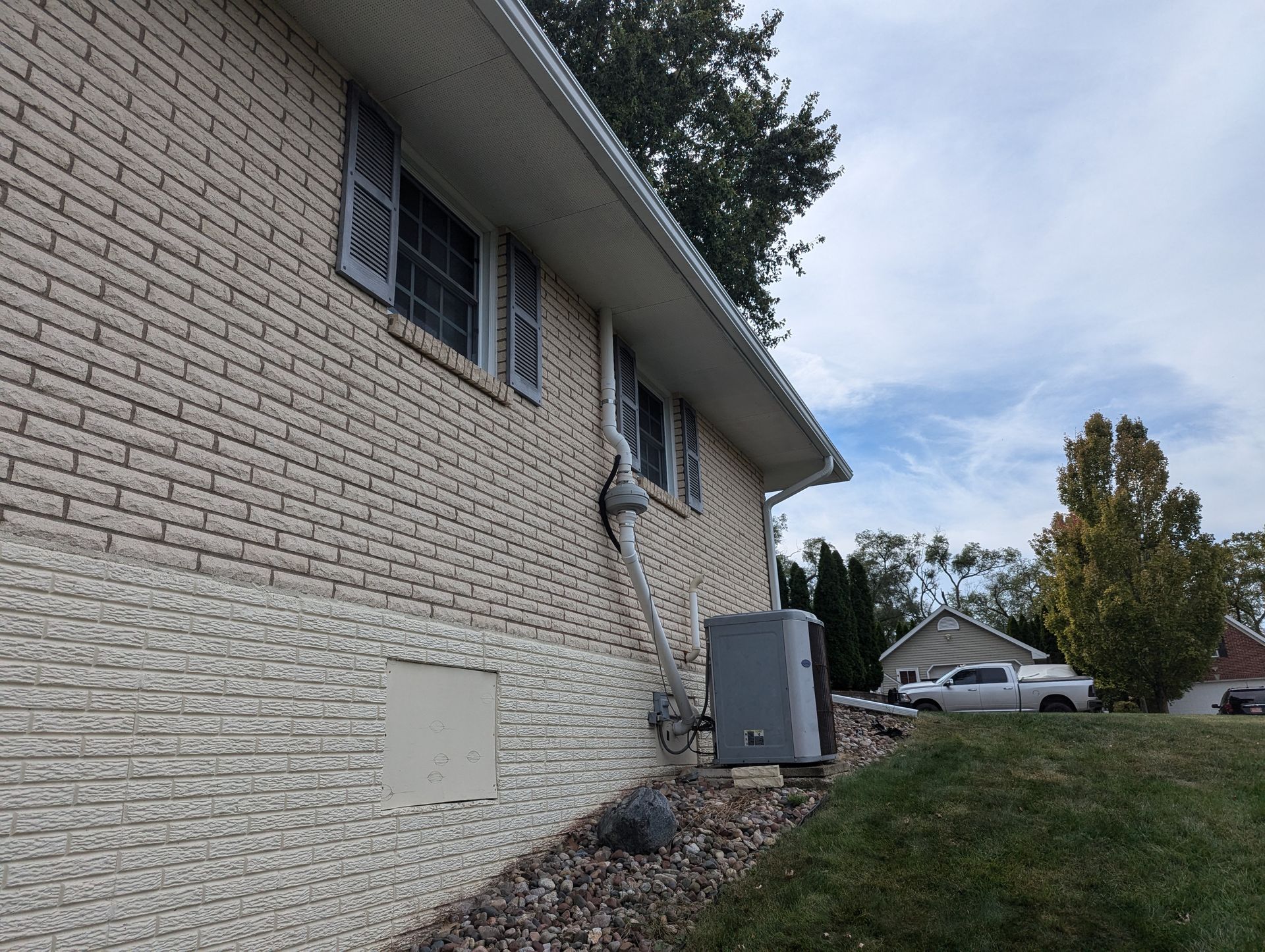 Side view of a brick house with gray trim, gutters, and an air conditioning unit on a grassy hill.