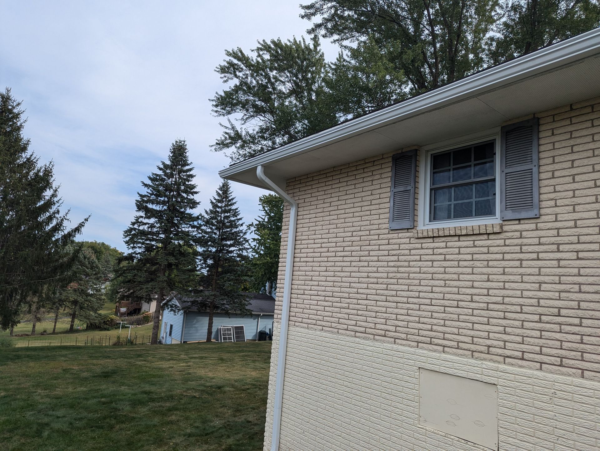 Side of a brick house with a white trim, gutters, and a window with gray shutters, trees and sky in the background.