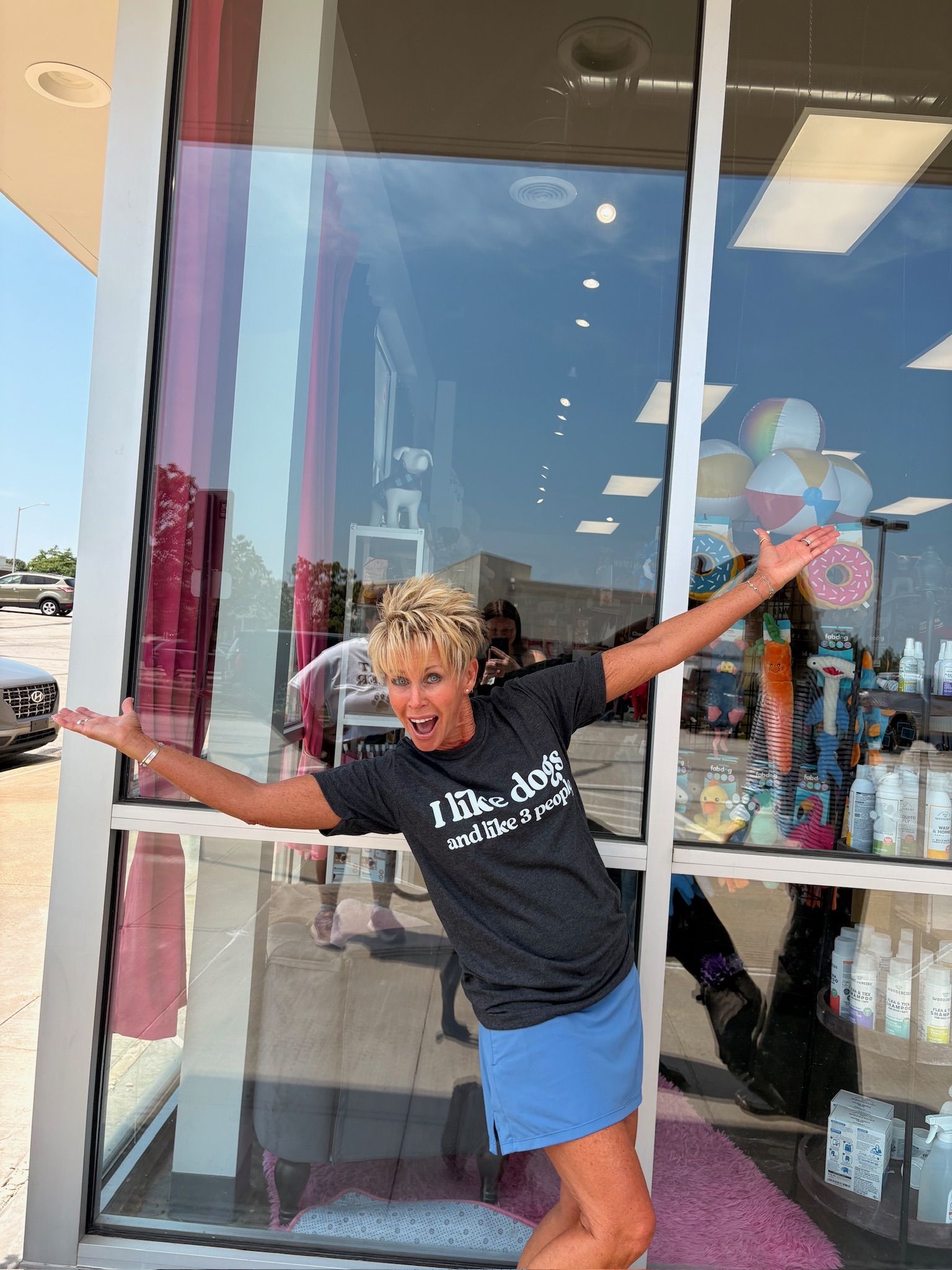 Person posing with arms outstretched in front of a storefront window, smiling and leaning playfully.