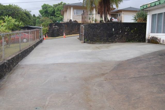Empty concrete driveway beside a stone wall and fence, with houses and a palm tree in the background