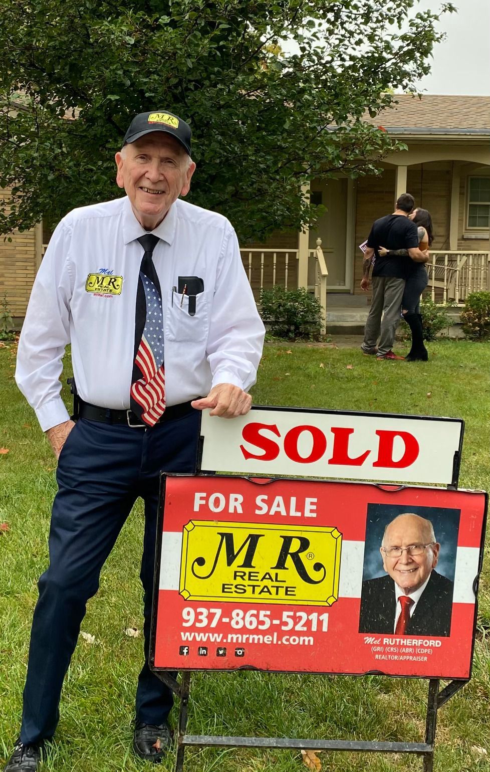 A man is standing in front of a house holding a sold sign.