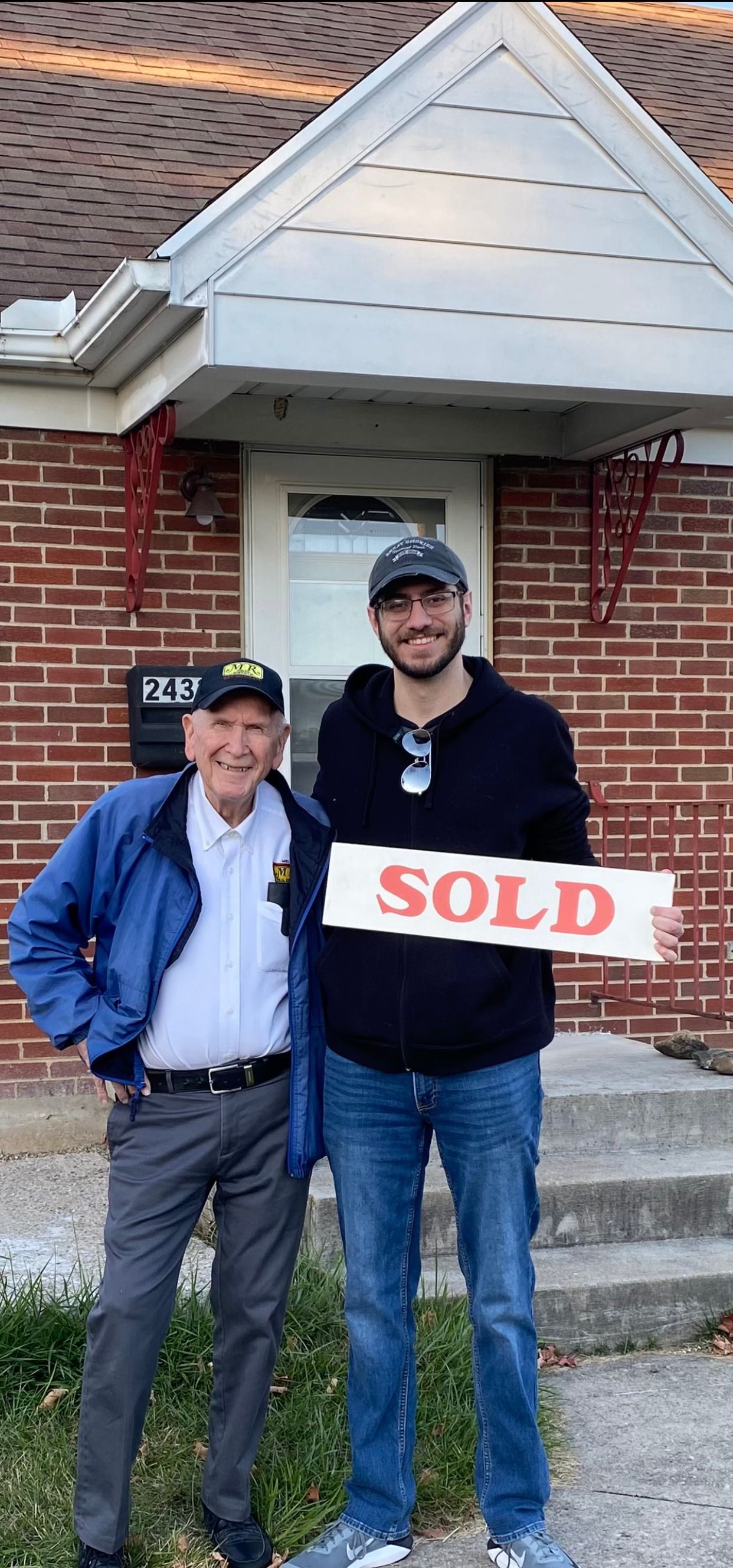 Two men are standing in front of a brick house holding a sold sign.