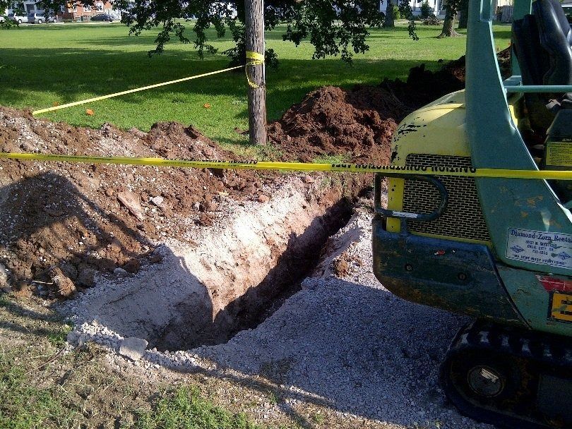 Construction excavator digging a trench beside a yellow caution tape in a park