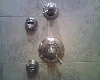 Chrome shower controls on a tiled bathroom wall, including a main handle and two smaller knobs.