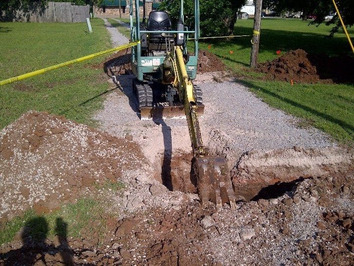Backhoe digging a trench in a gravel driveway beside a grassy yard