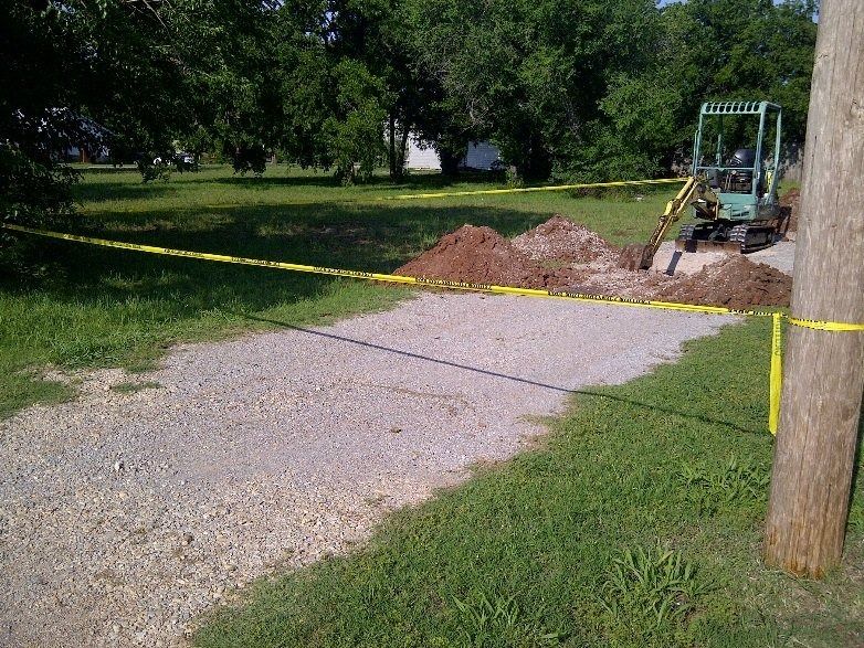 Yellow caution tape blocks a gravel path beside dirt piles and a small excavator in a grassy park.
