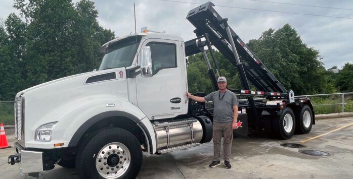 Man stands beside a white roll-off truck with raised container. Outdoors, daytime.