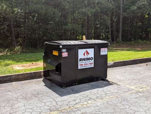 Black dumpster with "RHINO" logo on asphalt in front of trees.