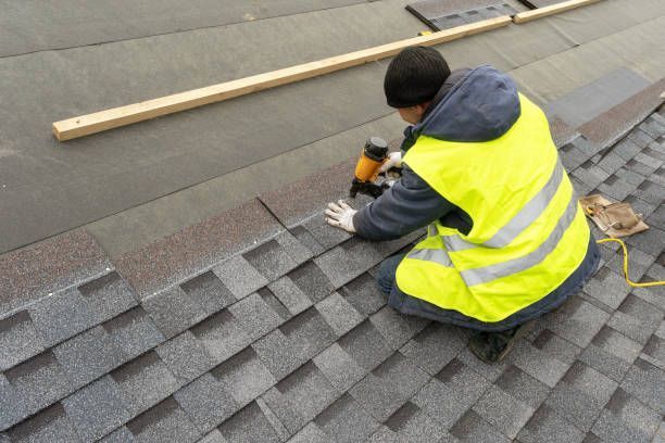 Roofer in safety vest kneeling on a roof, using a nail gun to install shingles.