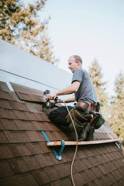 Roofer installing shingles on a roof. Brown shingles, gray shirt, tool belt, and sunny outdoor setting.