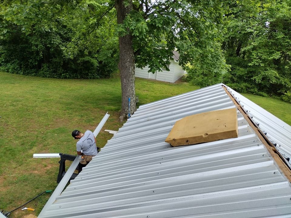 Man installing gutter on a metal roof, yellow insulation block, tree, and green grass.