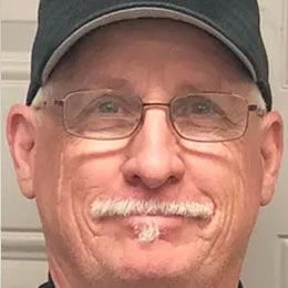 Close-up of a smiling man wearing glasses and a dark cap, with a white mustache.