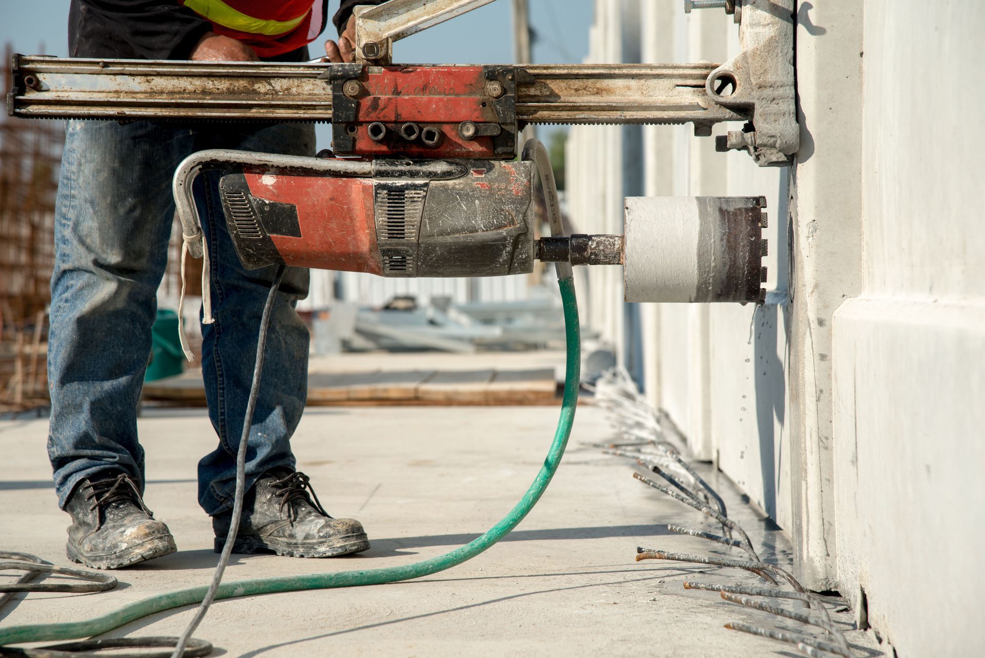 Worker using a core drill to bore a hole in a concrete wall, with green hose on a construction site