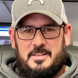 Man wearing glasses and a gray cap, seated indoors, looking at the camera with a slight smile