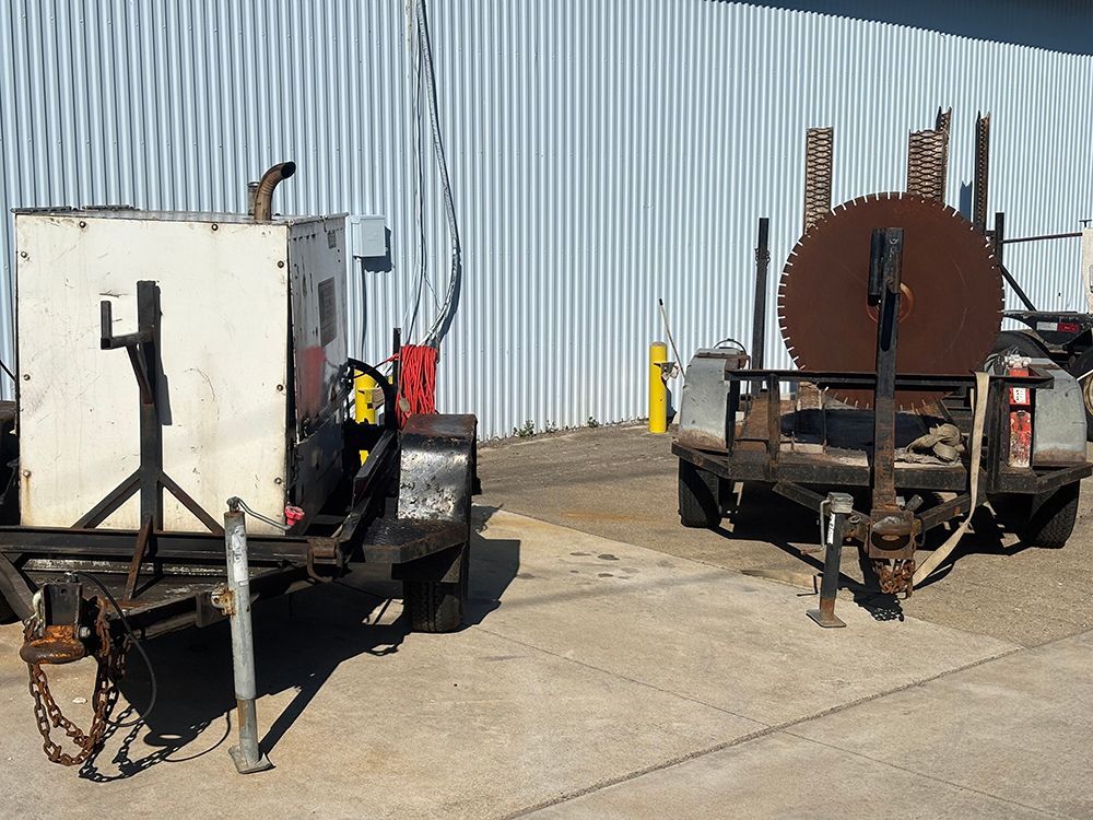 Two utility trailers parked on concrete beside a corrugated metal building, one with a tank and chains.
