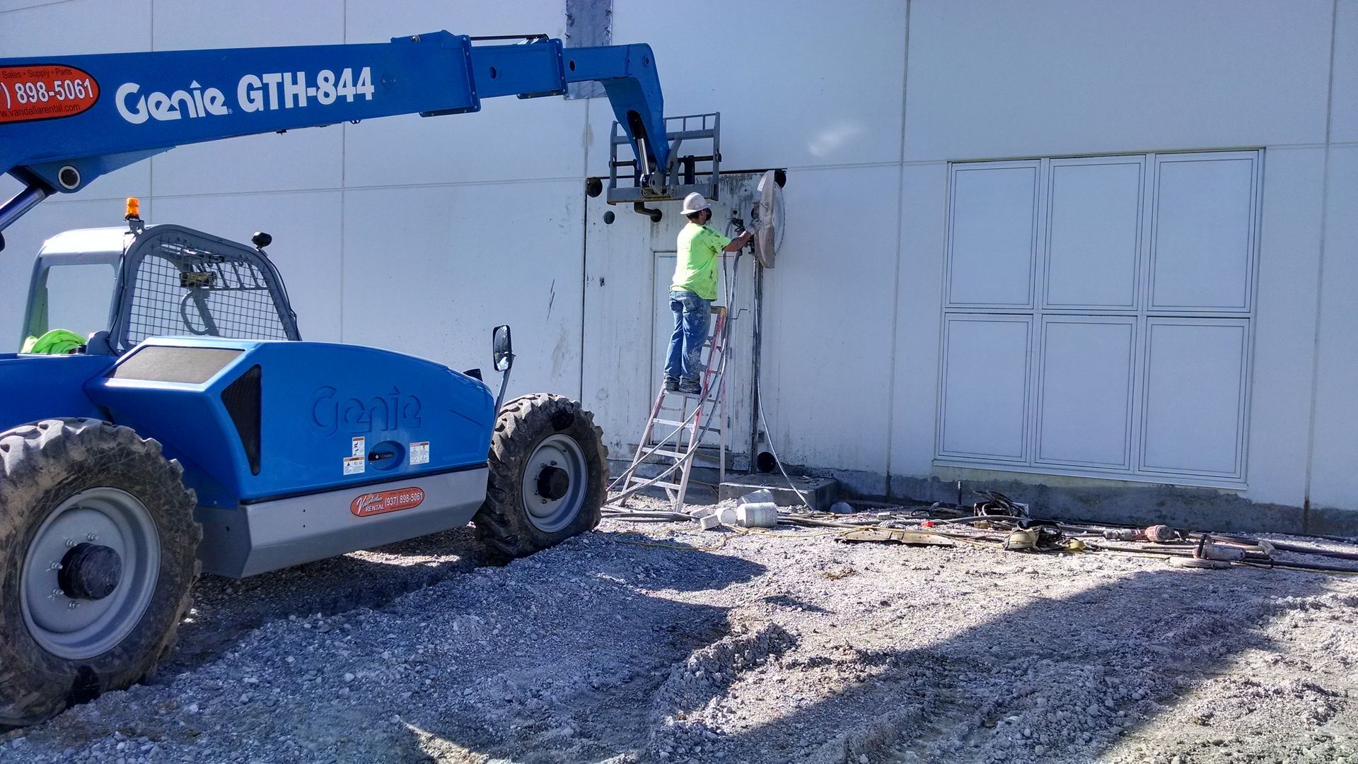 Blue boom lift with worker repairing a wall beside a white industrial building.