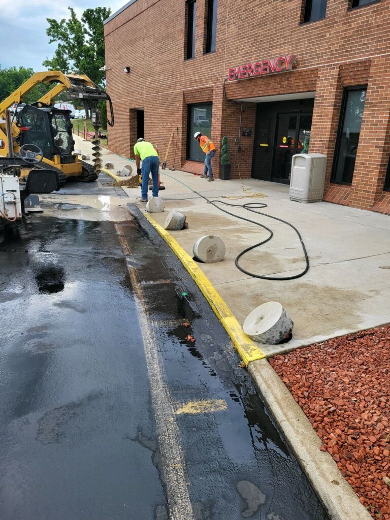 Workers repair a brick building entrance beside wet pavement, a backhoe, and hoses.