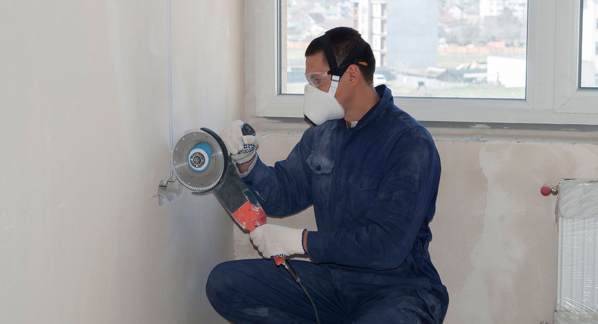 Person in a mask using an angle grinder to cut into a wall near a window in a room under renovation