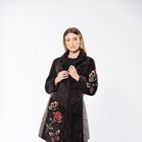 Woman wearing a dark coat with floral embroidery, hands near the front, posing against a white backdrop.