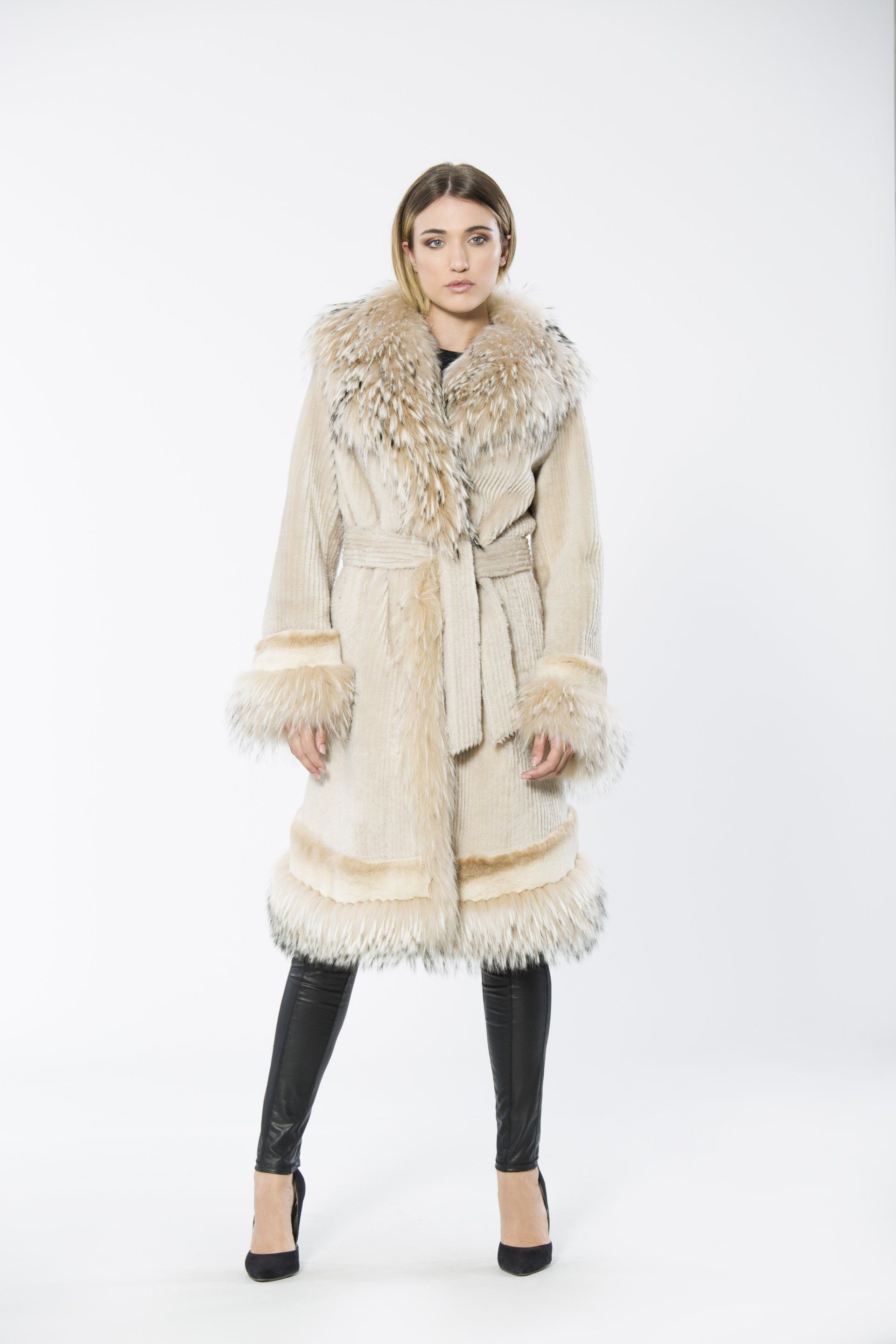 Woman in long beige fur coat, black leggings, and heels, standing against white background.